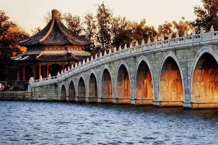 Elegant arched stone bridge beside a traditional Chinese pavilion over a serene lake during sunset in China.