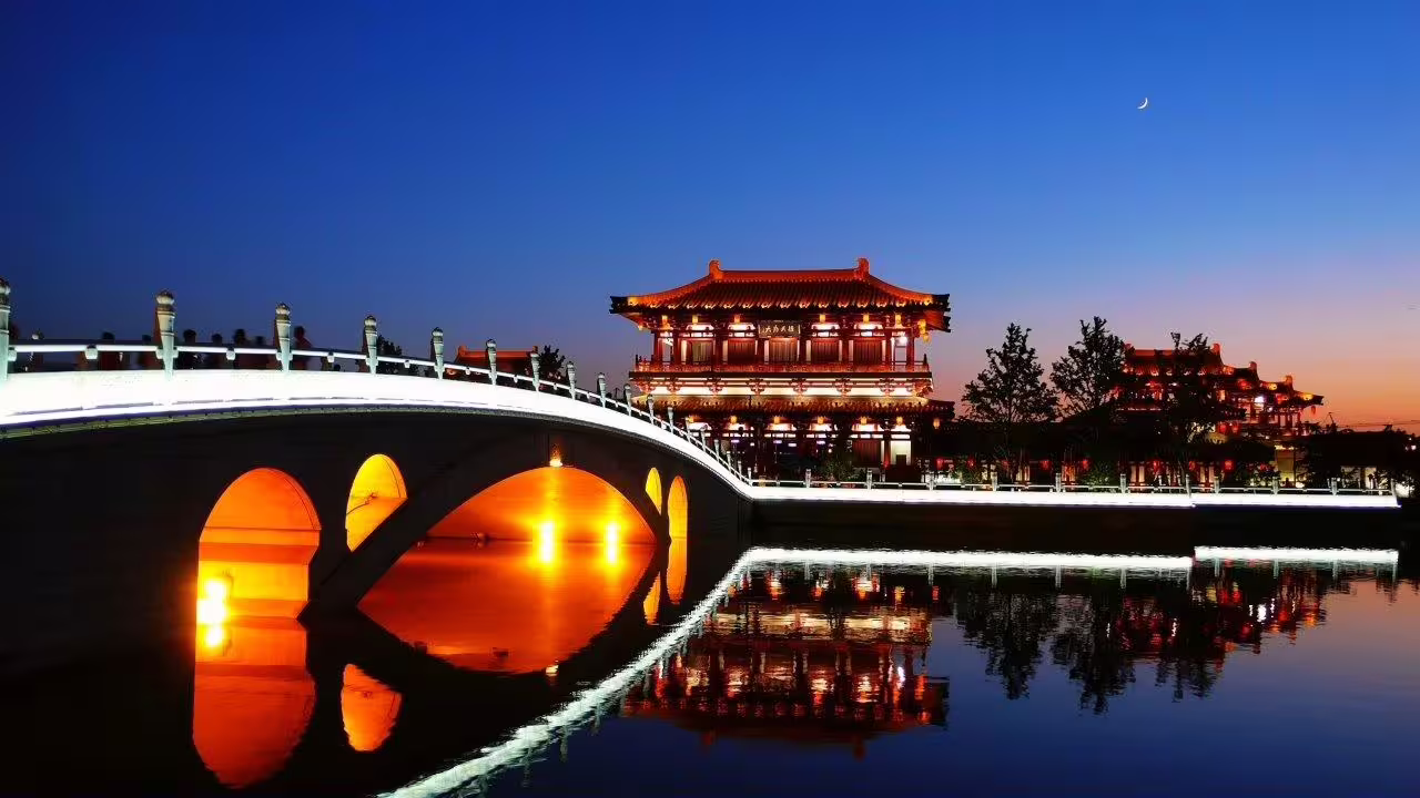 Discover a beautifully illuminated Chinese bridge and pavilion reflecting in a tranquil river at dusk on the Mysterious West of China tour.