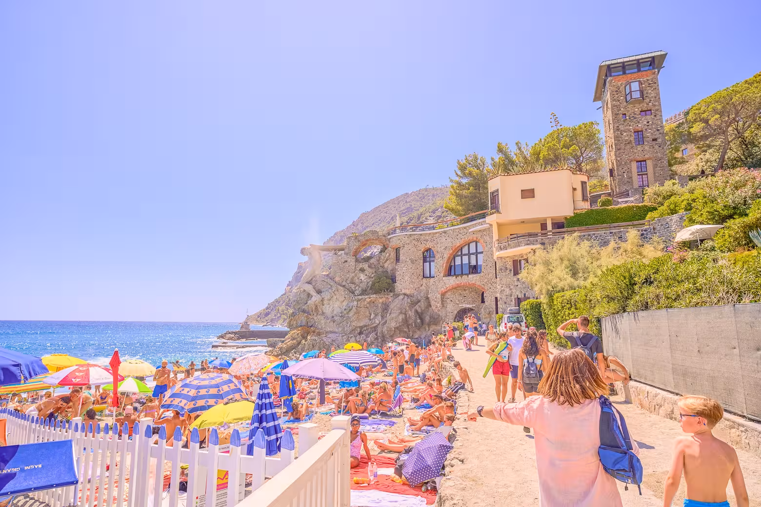 Bustling beach scene in Cinque Terre with vibrant umbrellas, sunbathers, and historic cliffside architecture.