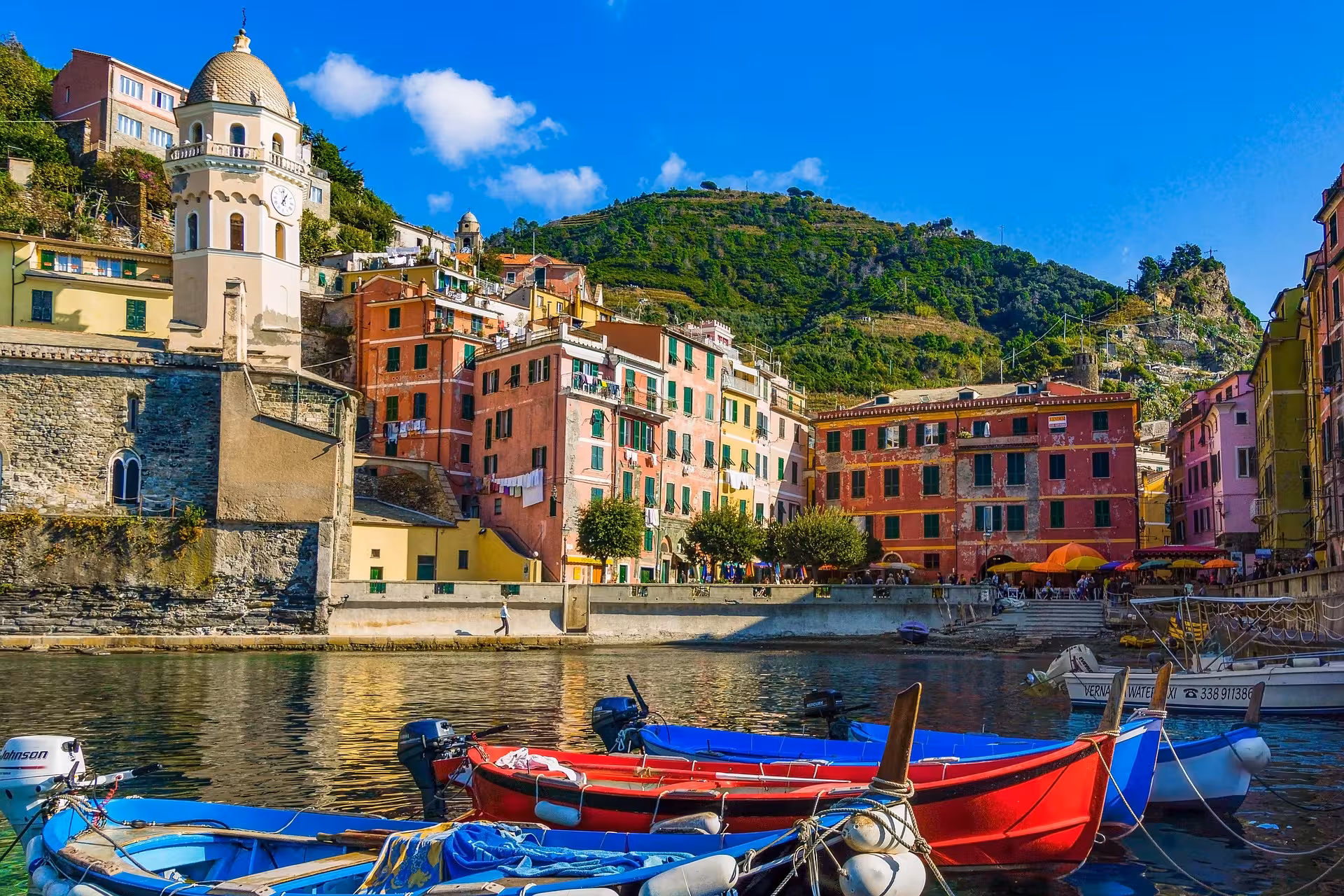 Colorful boats docked in Vernazza harbor with vibrant hillside buildings and lush greenery, Cinque Terre boat tour.