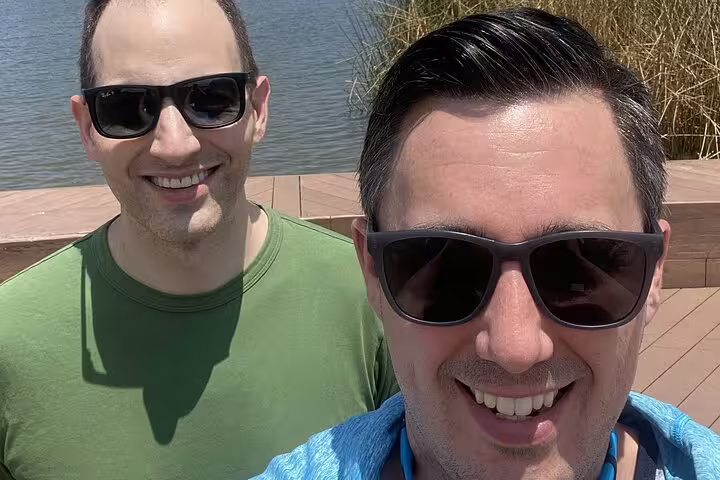 Two smiling men wearing sunglasses taking a selfie outdoors near a waterway on a sunny day