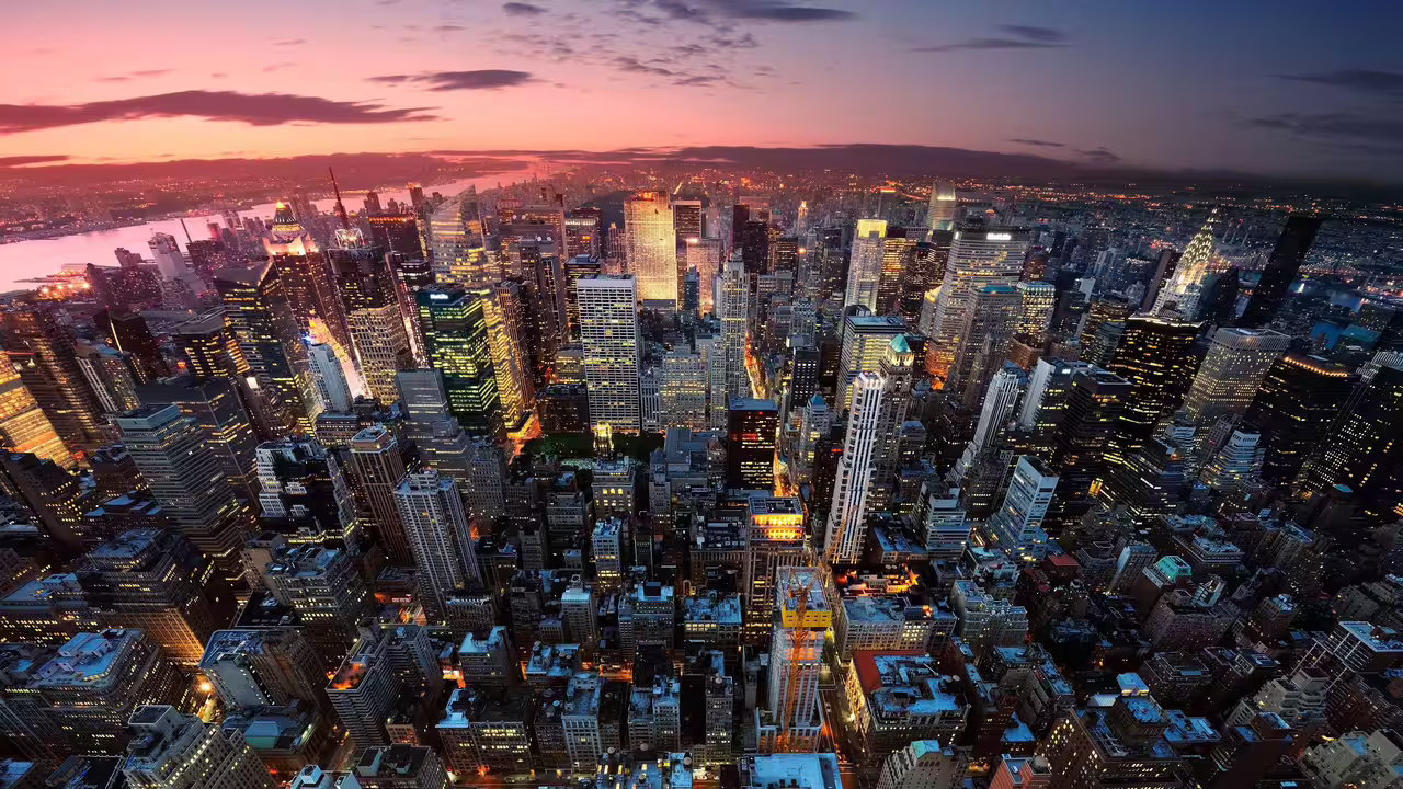 Aerial view of a vibrant city skyline at dusk, showcasing illuminated skyscrapers and a colorful sunset backdrop.