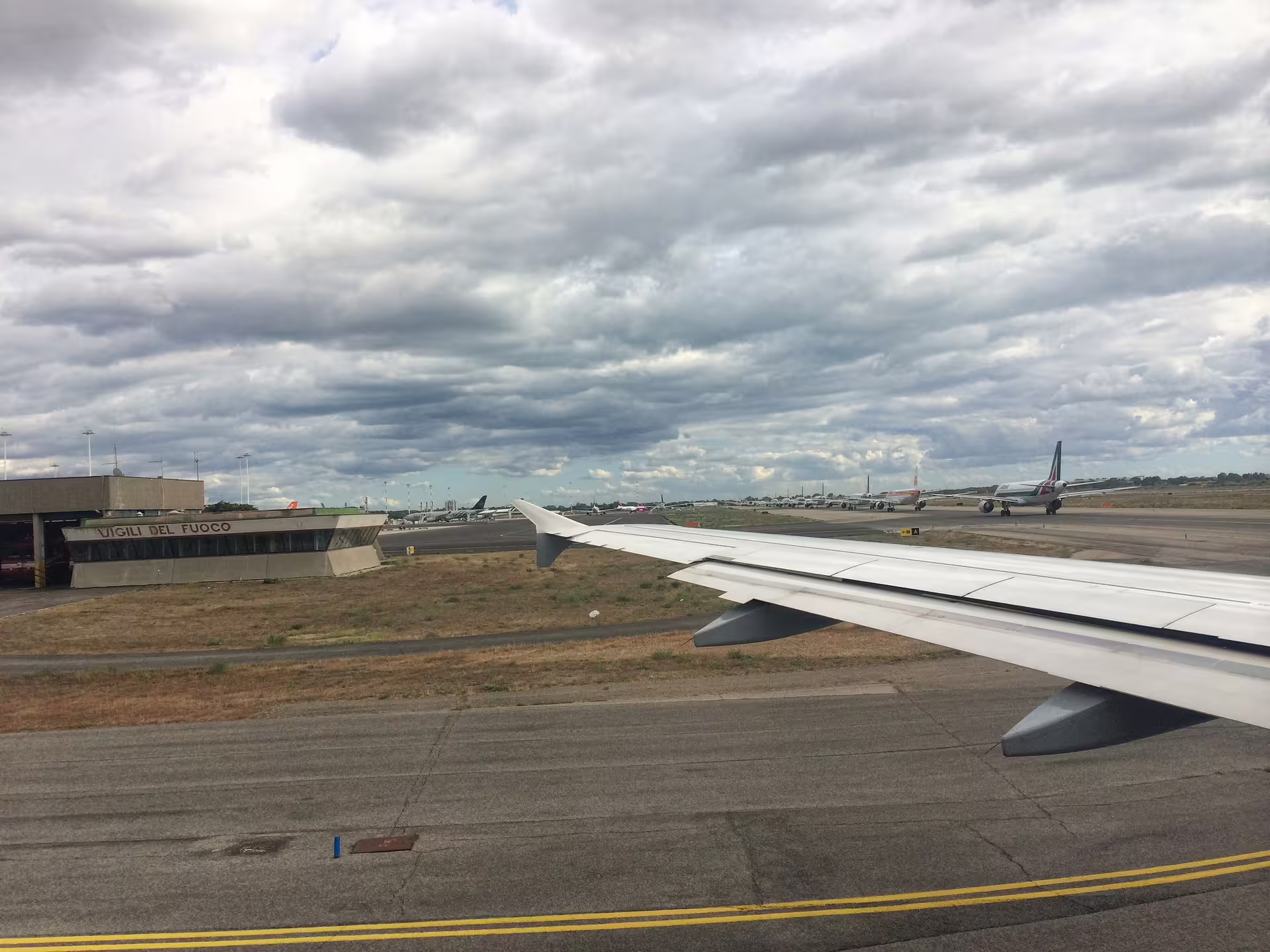 Passenger view of aircraft wing taxiing on runway at Rome Fiumicino Airport under cloudy sky before cruise transfer
