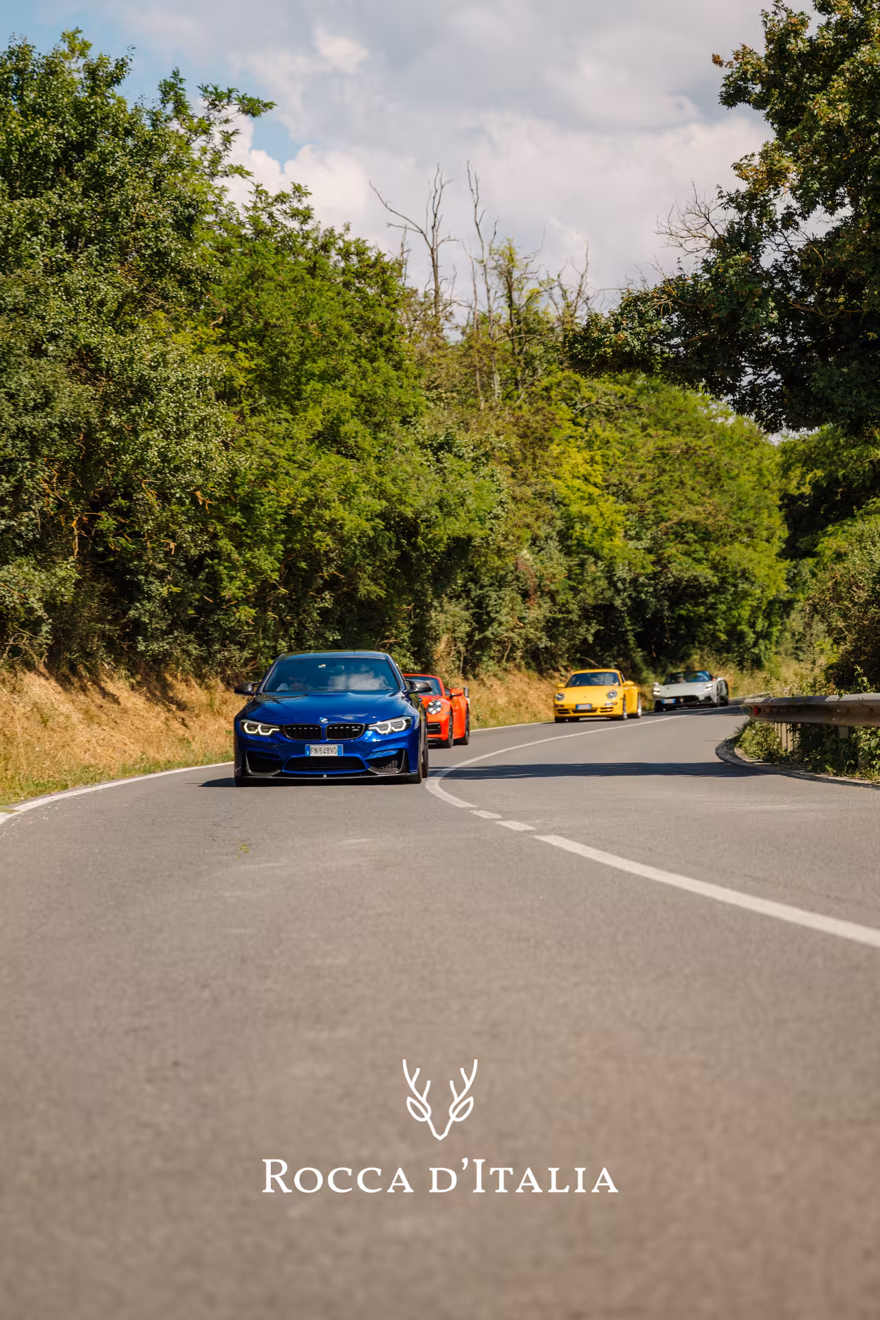 Convoy of sports cars led by blue BMW M4 on a scenic tree-lined road in Chianti, Italy