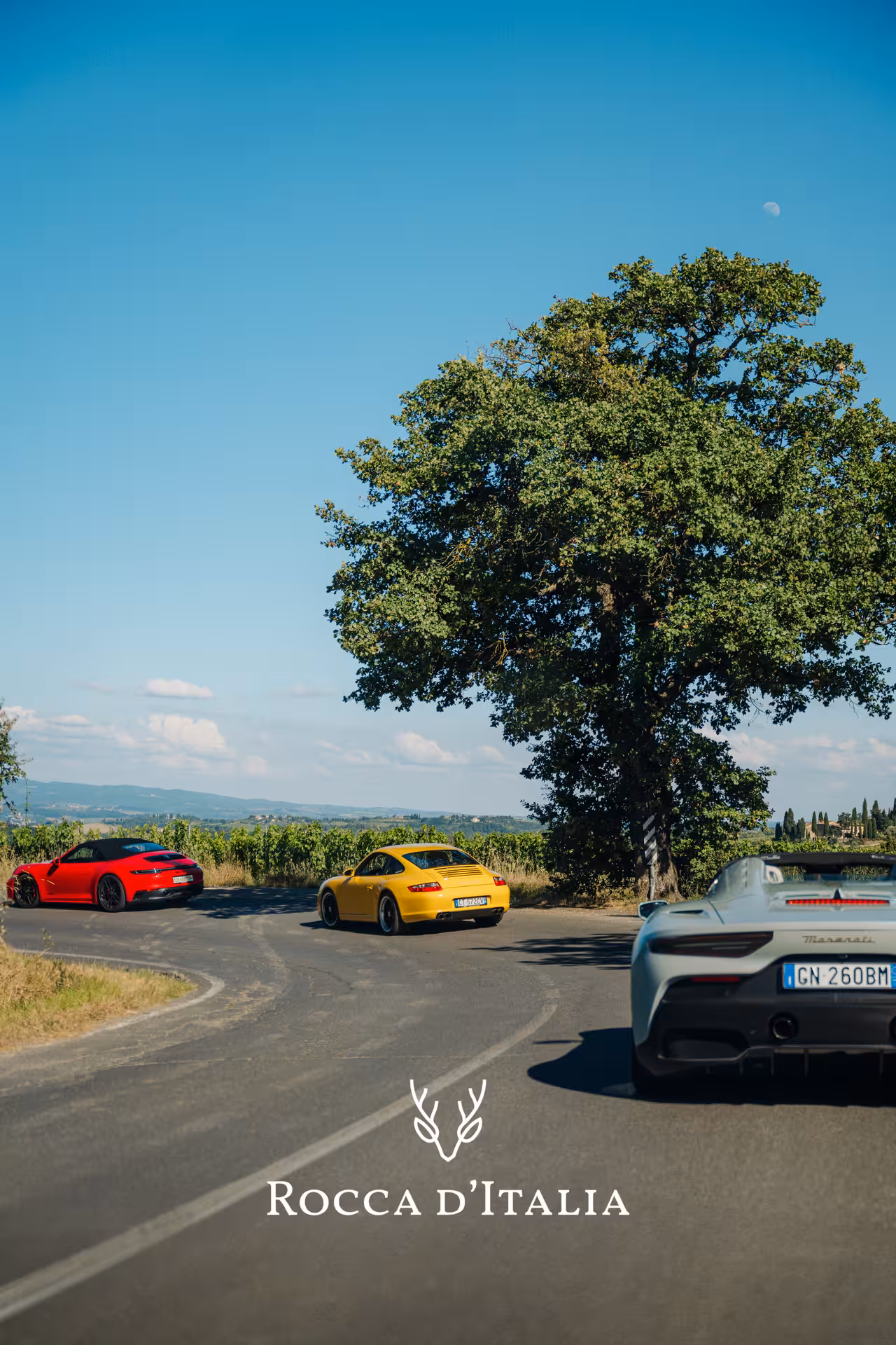 Three sports cars including a Maserati and Porsches driving through Chianti vineyards on a sunny day