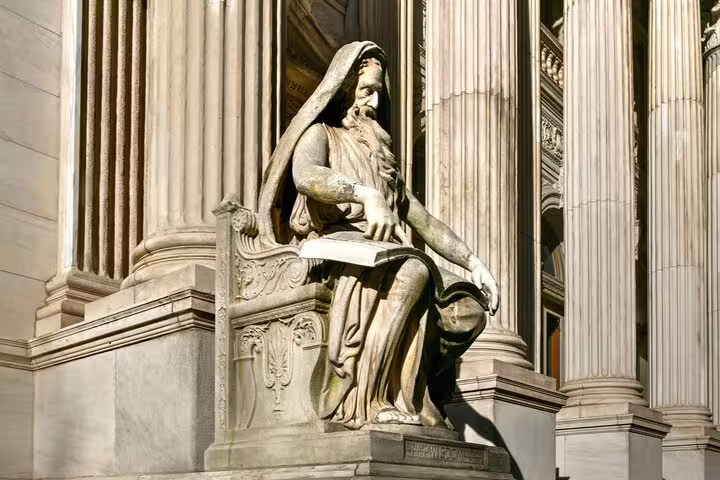 Classical marble statue of a robed figure reading a book, seated before tall stone columns