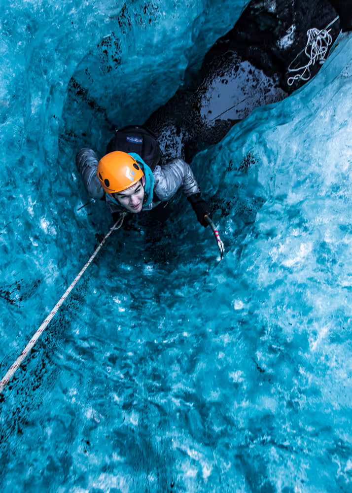 Climber in orange helmet ascending a stunning blue ice cave wall with rope on a private glacier climbing photoshoot adventure