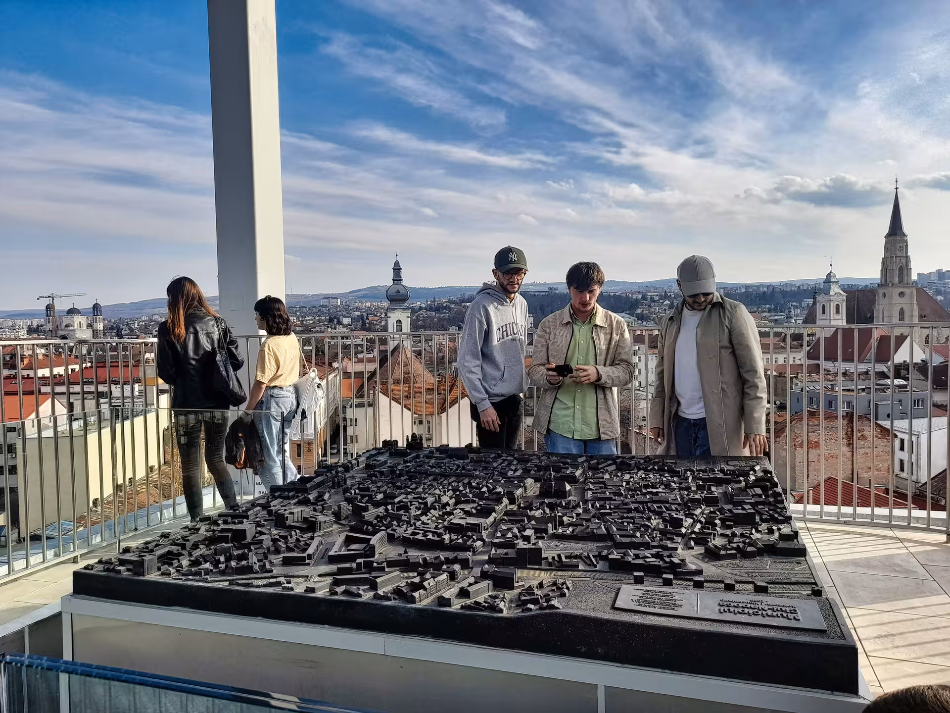 Tourists exploring a detailed Cluj-Napoca city scale model on a rooftop with panoramic views of historic churches and skyline during a guided walking tour.