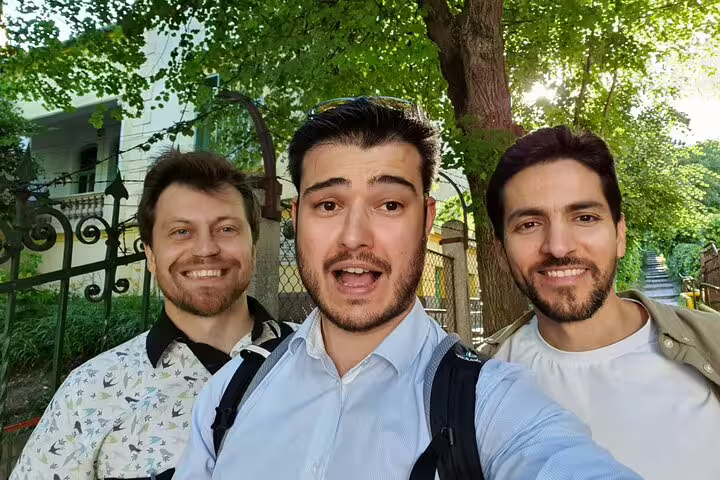 Three smiling men taking a selfie on a walking tour in Cluj-Napoca, with green trees and iron gate behind them