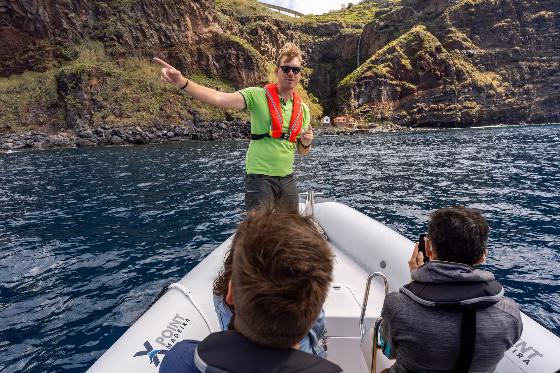 Guide points out scenic coastal landmarks to tourists on a RIB boat tour near rocky cliffs.
