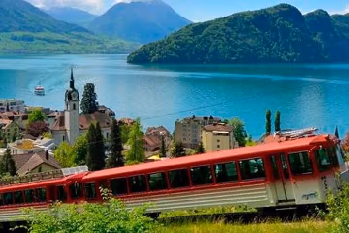 Scenic shot of a red cogwheel train passing by a lakeside village with Lake Lucerne and mountains in the background.