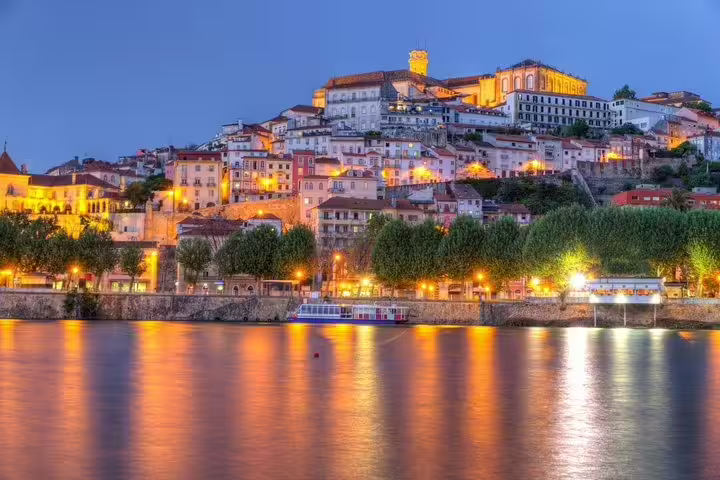 Scenic evening view of Coimbra's historic hilltop architecture reflecting in the Mondego River, perfect for a day trip from Lisbon.