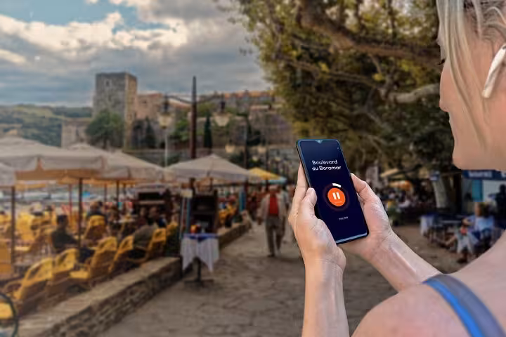 Woman holding smartphone with GPS audio guide app on Collioure waterfront promenade with castle in background