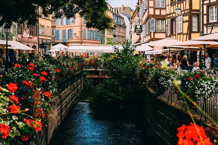 Flower-lined canal with colorful half-timbered buildings and outdoor cafes in Colmar, Alsace