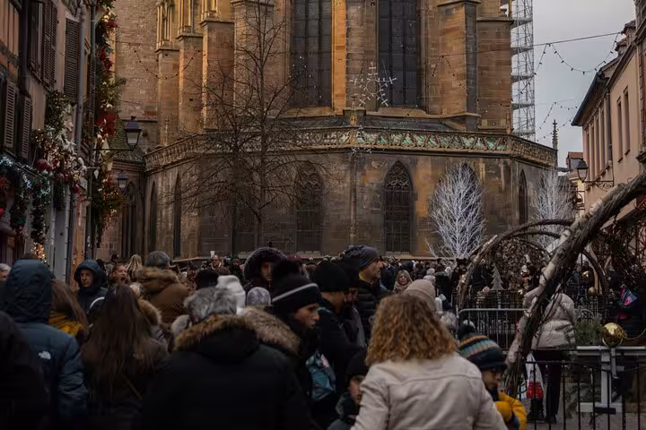 Crowds exploring Colmar Christmas market with historic Gothic church in background, Alsace France