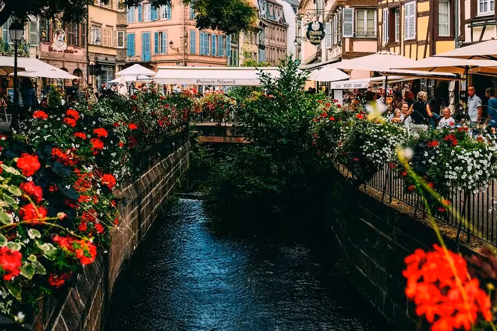 Flower-lined canal in Colmar old town with colorful half-timbered buildings and outdoor cafés