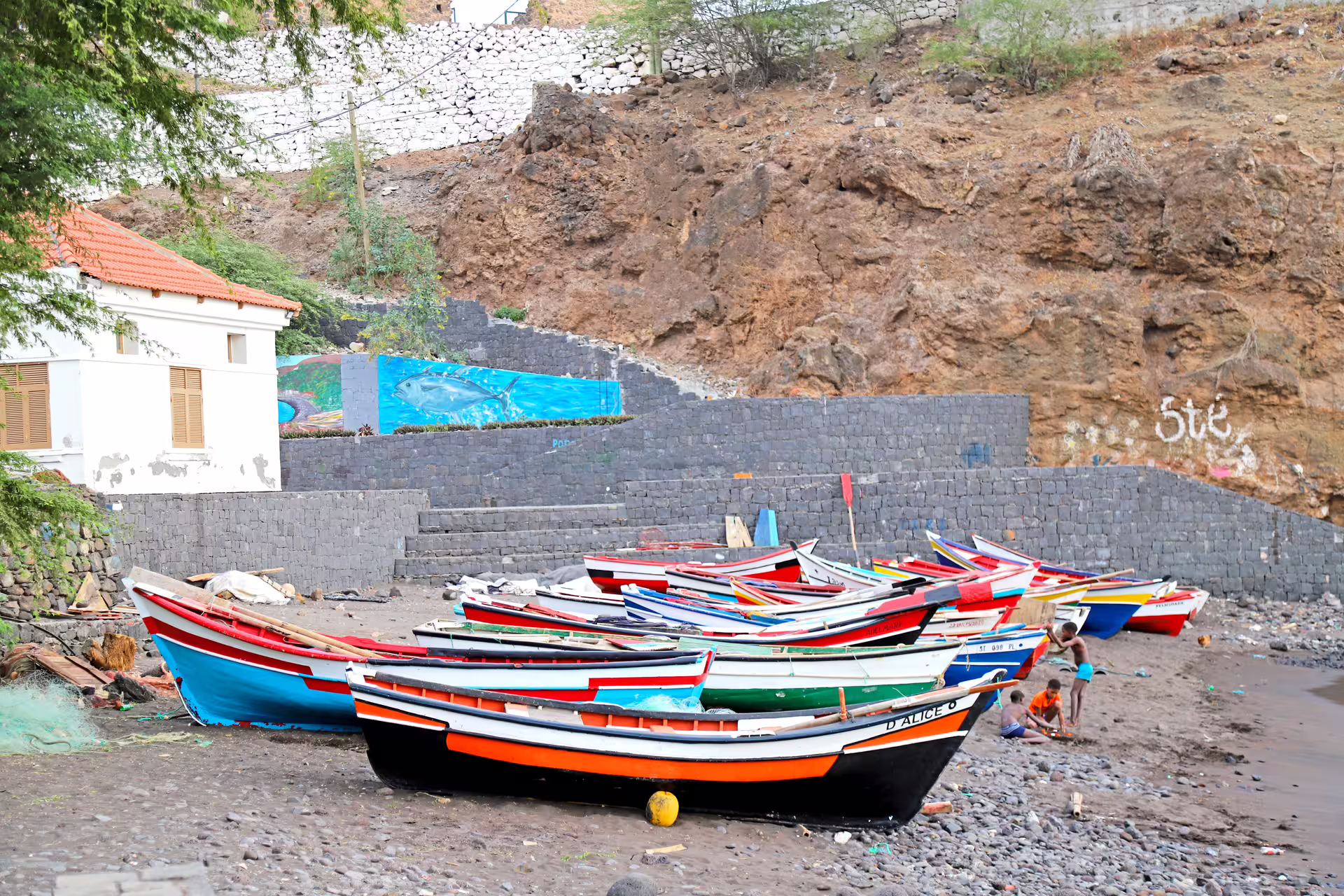 Colorful fishing boats on Cidade Velha's beach with stone walls and mural backdrop.