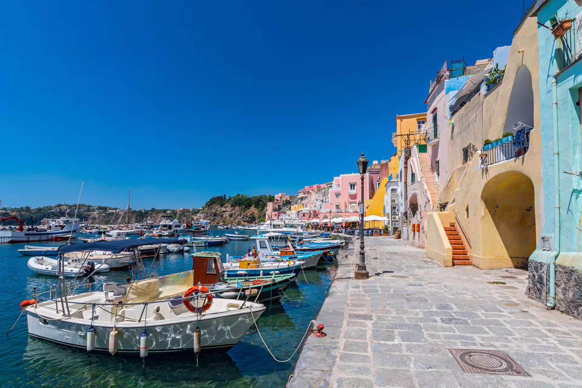 Colorful fishing boats moored along Procida harbor with pastel houses and clear blue sky on Ischia & Procida boat tour from Sorrento