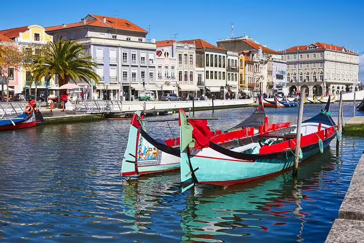 Colorful moliceiro boats docked on Aveiro's canal, with vibrant historic buildings and palm tree-lined streets in the background.
