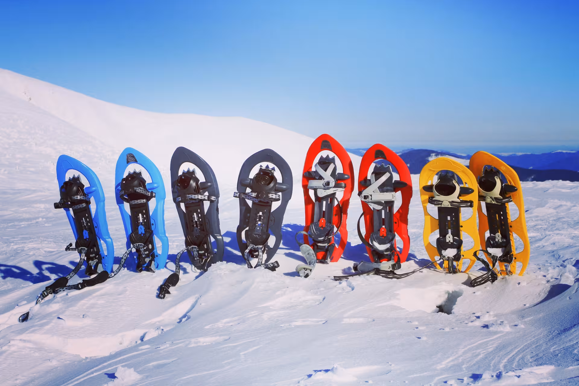 Colorful snowshoes in blue, grey, red, and yellow lined up on a snowy mountain summit under clear blue sky