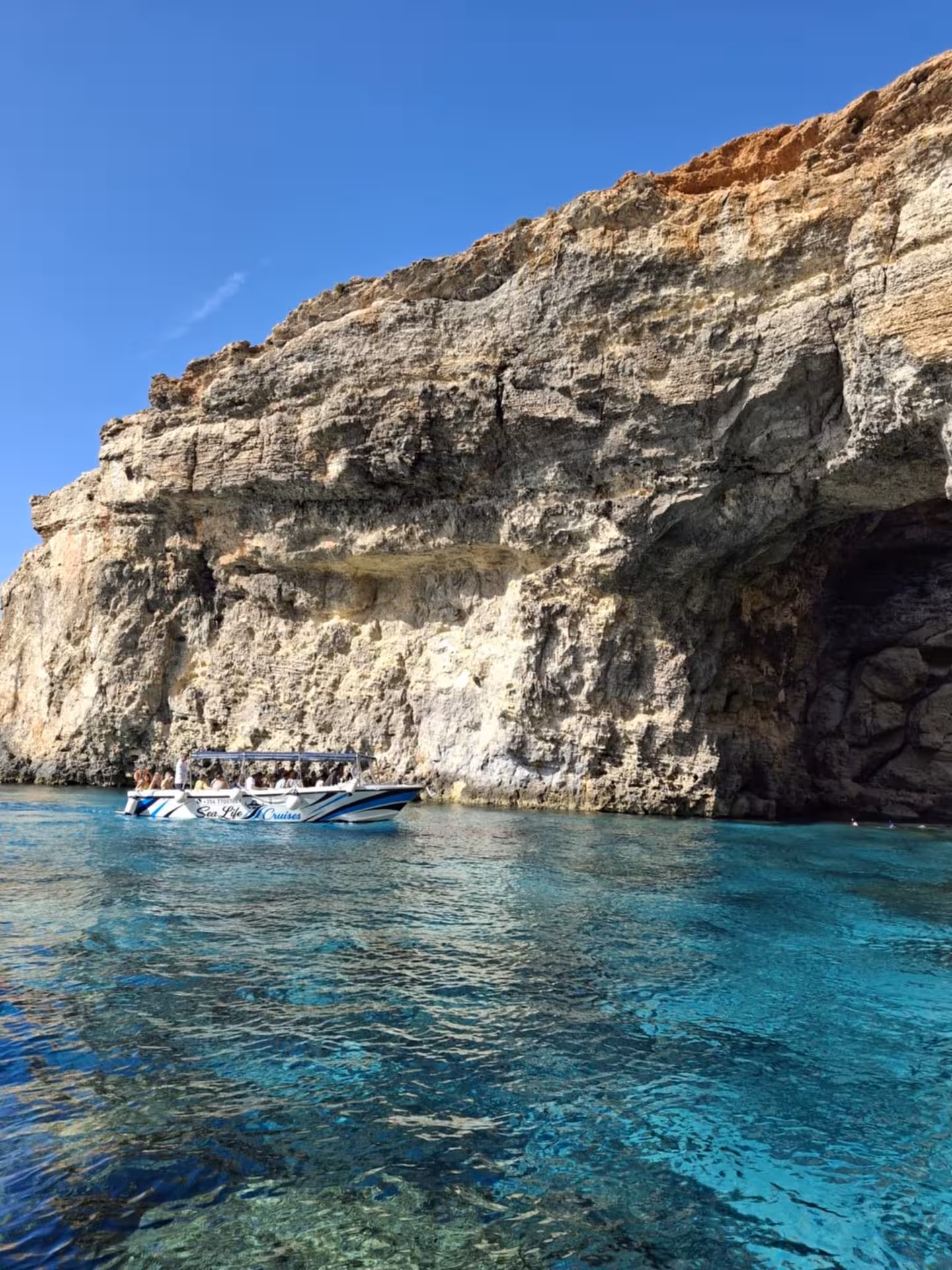 Comino sea caves cruise boat by towering cliffs in Crystal Lagoon, Malta, on Blue Lagoon evening tour