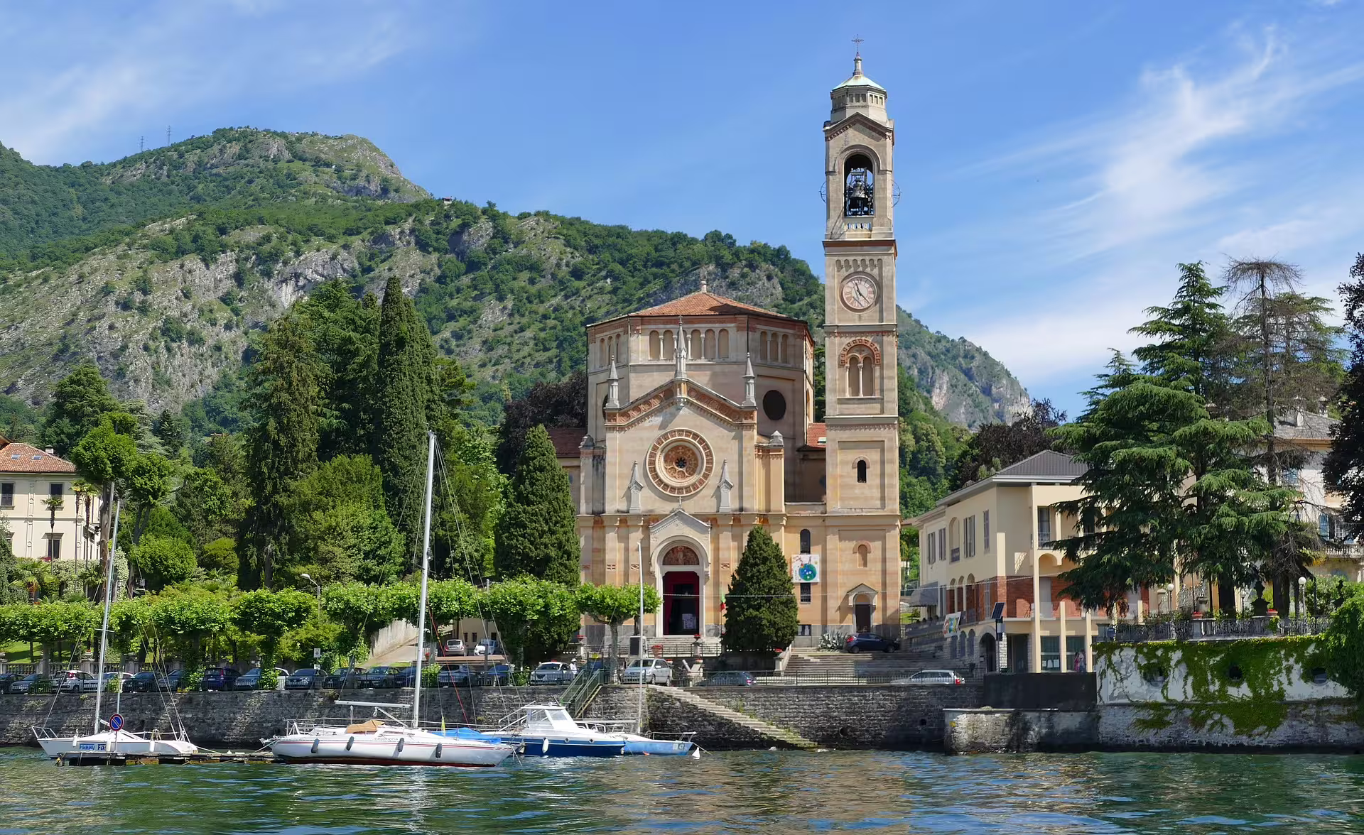 Historic lakeside church and bell tower in Como with sailboats, green hills and blue sky seen on a 2 hours walking tour