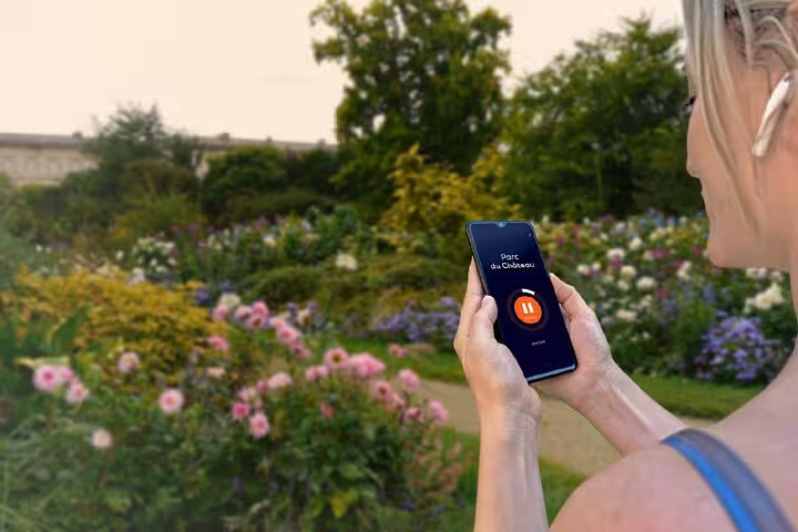 Woman holding smartphone with Parc du Château audio guide app in a colorful garden in Compiègne