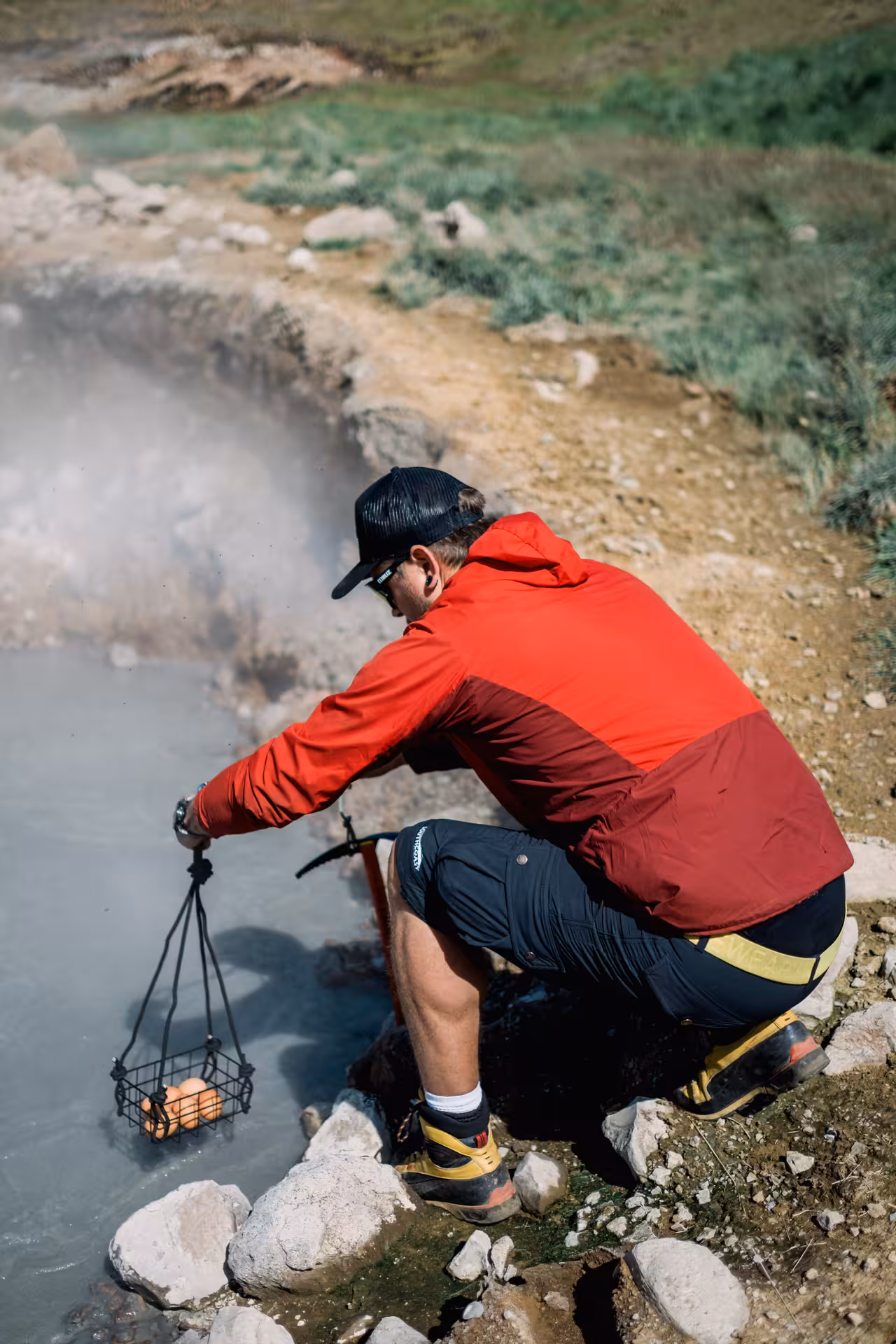 Hiker lowering eggs in wire basket into steaming geothermal hot spring in Iceland