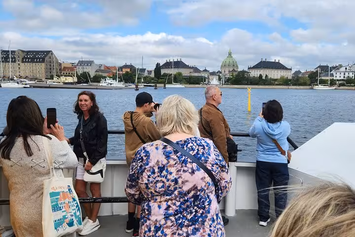 Tourists enjoy scenic waterfront views of Copenhagen landmarks from a ferry on a cloudy day.