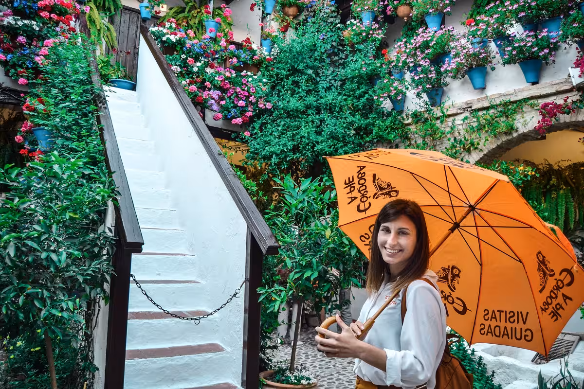Tourist enjoying a guided tour under a bright umbrella amidst lush greenery and flower-filled terraces in Córdoba.