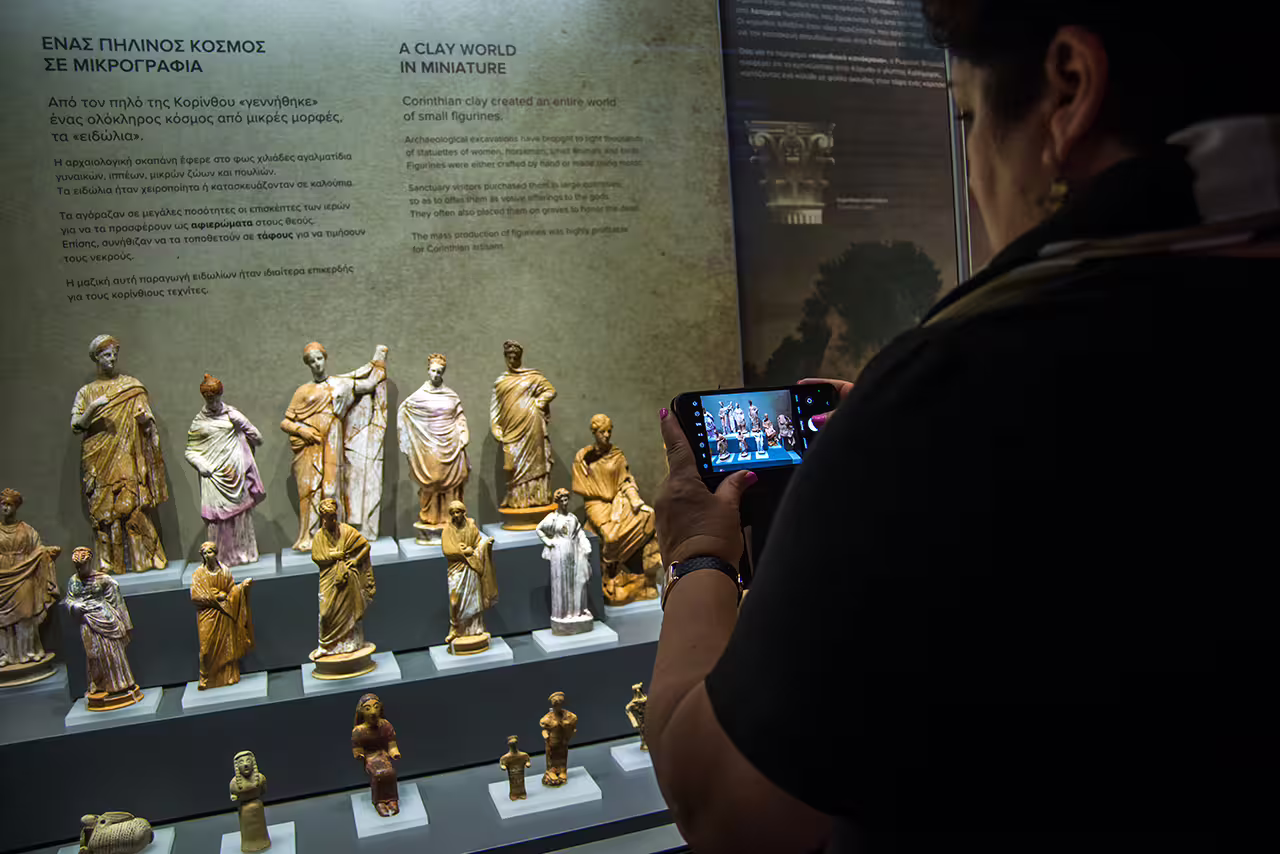 Visitor photographing Corinth Museum clay figurines exhibit on Ancient Corinth and Nafplion full-day guided tour