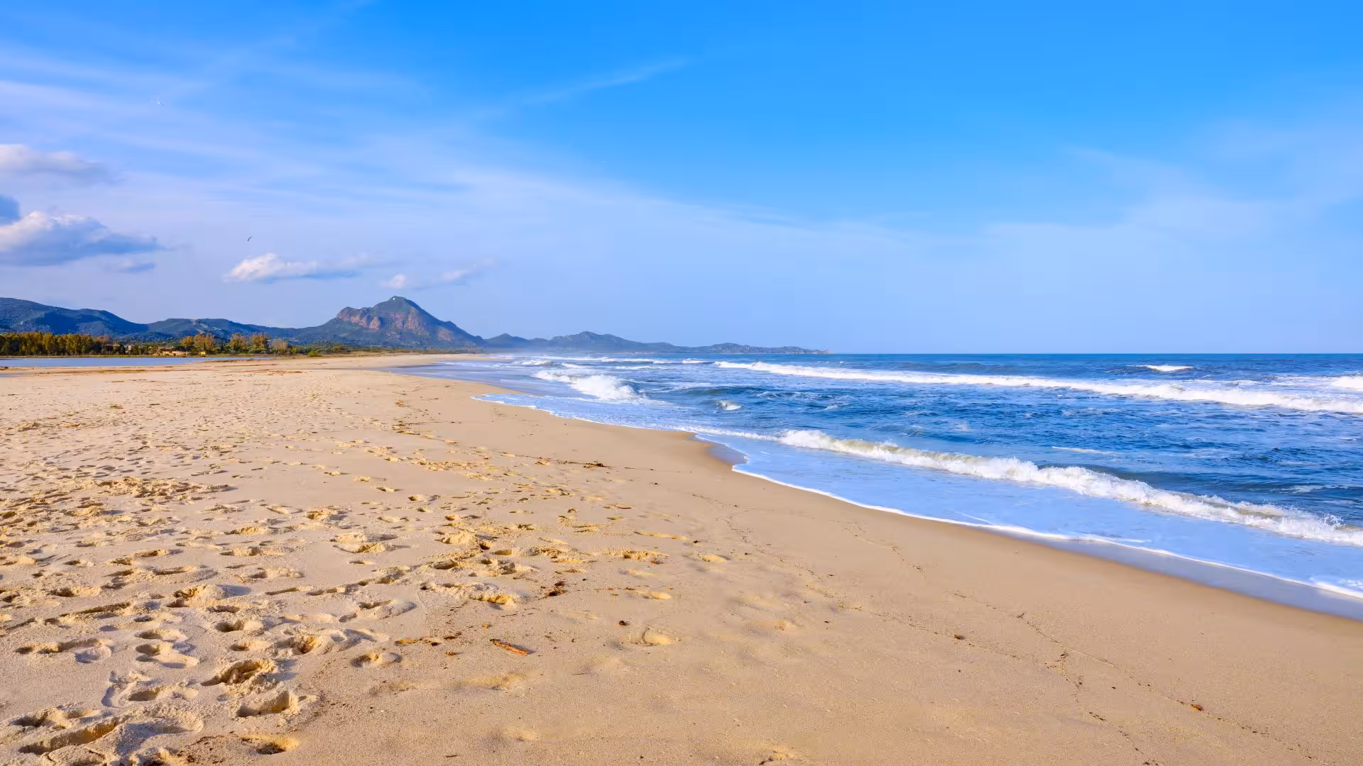 Expansive sandy beach at Costa Rei with gentle waves and distant mountains under a clear blue sky.