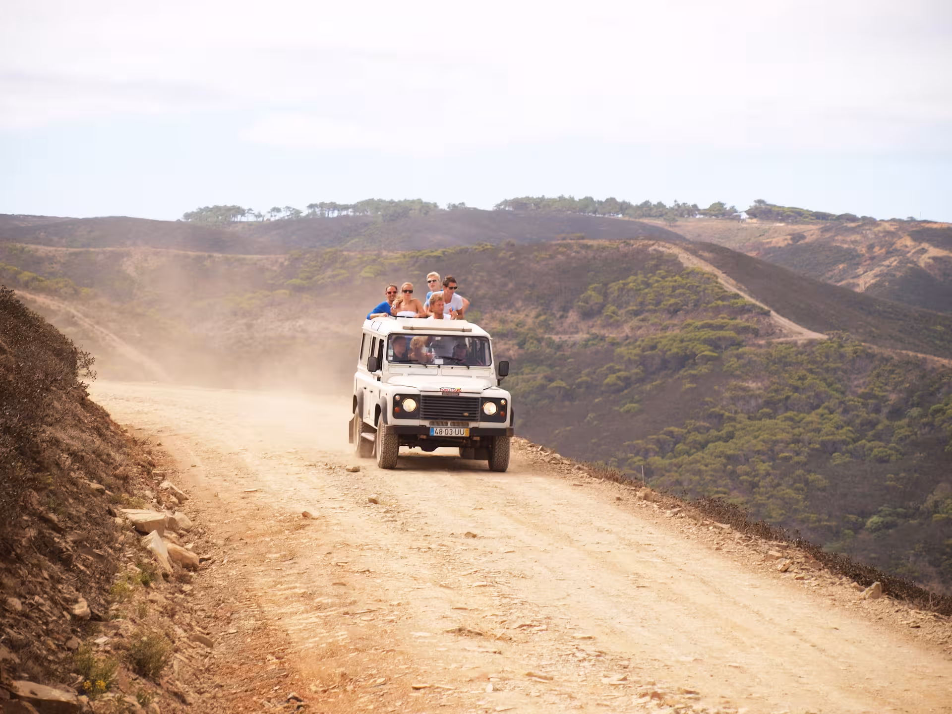 Open-top 4x4 jeep safari with smiling travelers driving a dusty cliff road in Costa Vicentina’s rugged hills