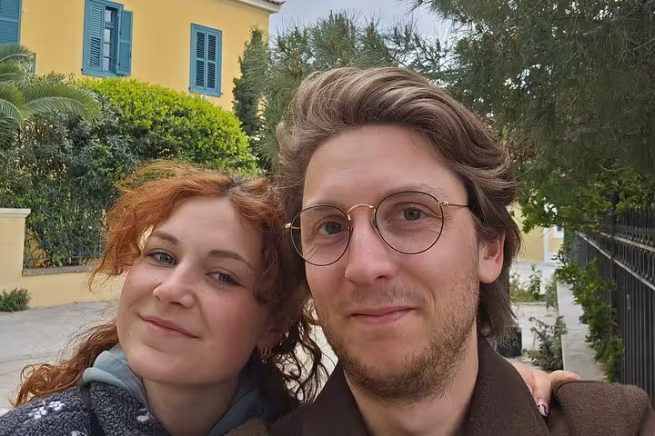 Couple smiling during self-guided Athens walking tour with yellow building and greenery in background