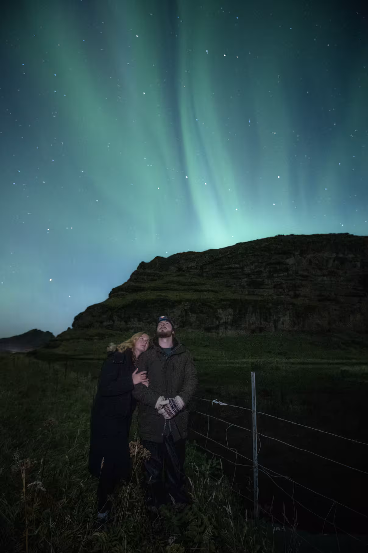 A couple embraces while gazing at vibrant green aurora borealis dancing over an Icelandic hillside on a magical northern lights dinner tour