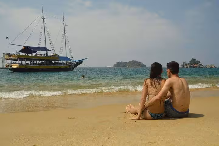 Couple relaxes on a sandy beach, watching a tour boat sail by in serene turquoise waters, perfect for a romantic getaway.
