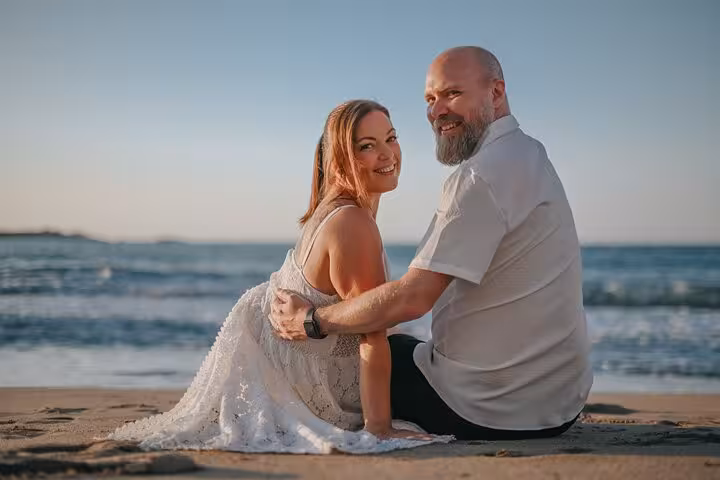 Smiling couple seated on a sandy beach in Hersonissos, enjoying a serene ocean view during a private photoshoot.
