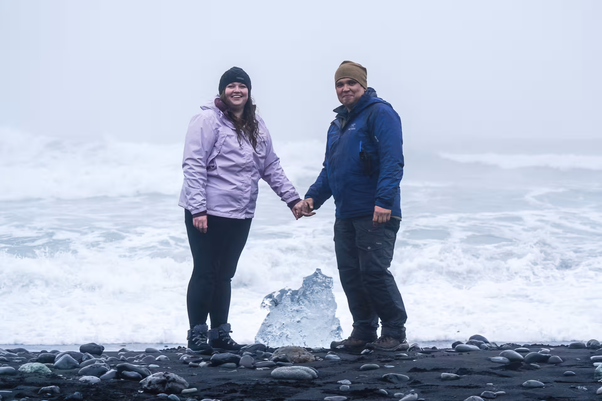 Couple holding hands on Iceland black sand beach with glacier ice chunk and crashing waves behind them