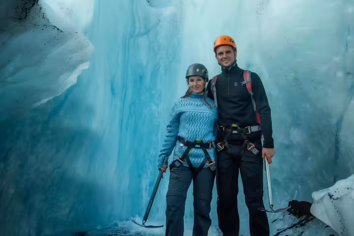 A smiling couple in helmets and harnesses pose inside a breathtaking blue ice cave on an Eggtrip Glacier Hike adventure.