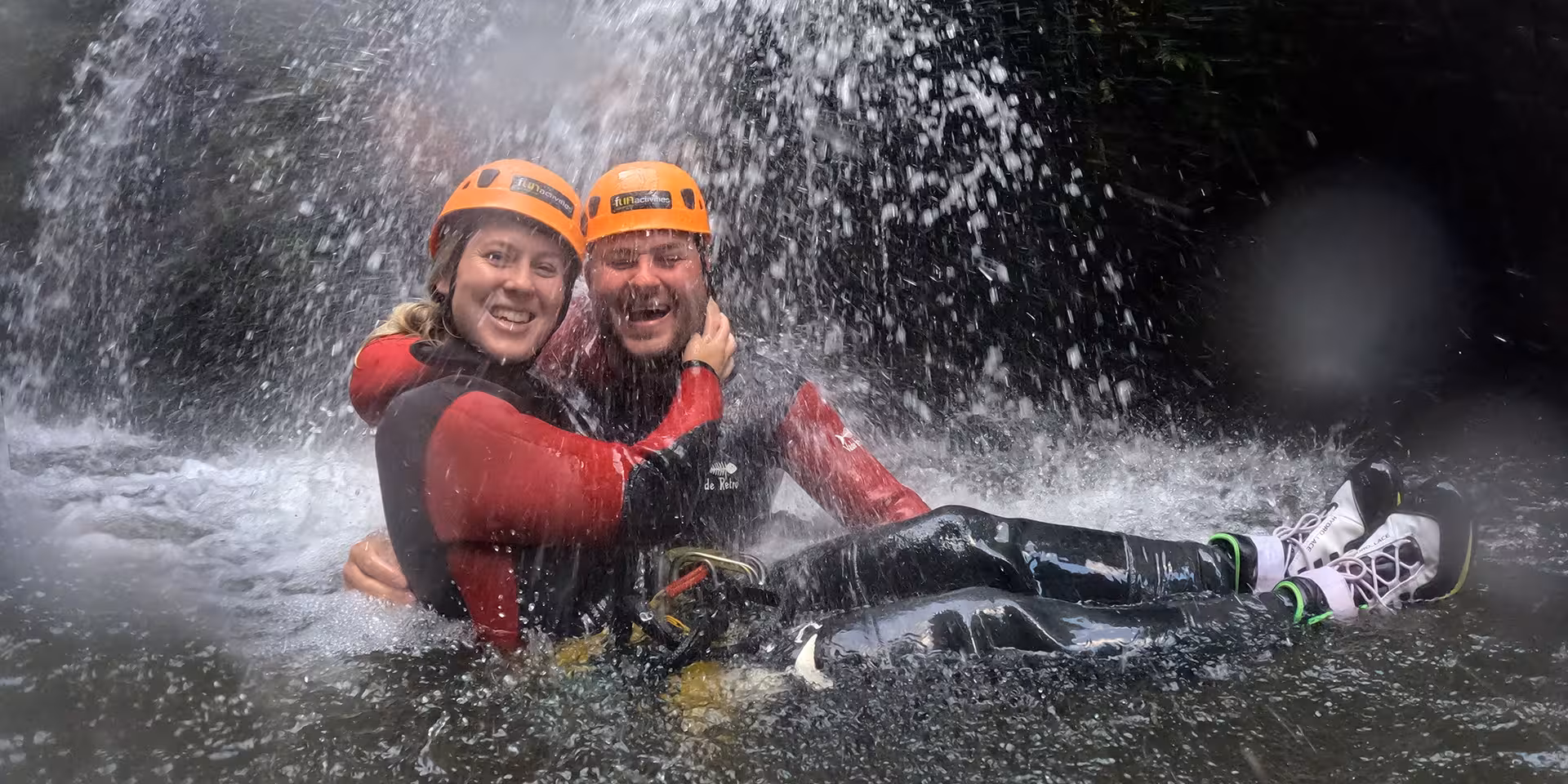 Happy couple canyoning under a waterfall in helmets and wetsuits, splashing in a natural pool adventure