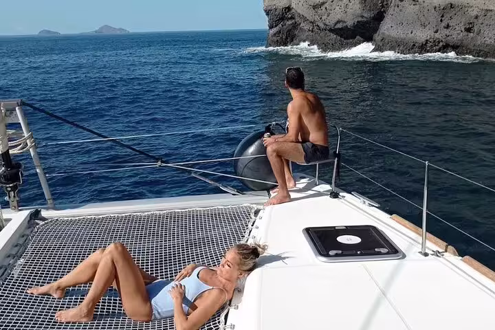 Couple relaxing on a catamaran with stunning views of Santorini's cliffs and deep blue sea.