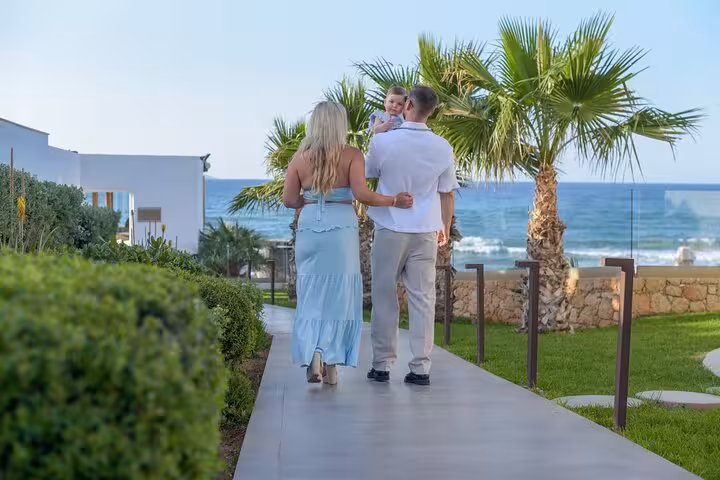 A couple walking hand in hand with their child along a scenic path bordered by palm trees and overlooking the sea in Analipsi.