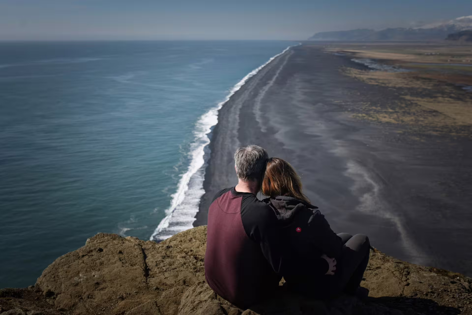 Couple sitting on cliffside overlooking Iceland's stunning black sand beach on a private South Coast tour