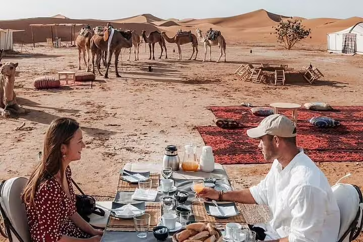 Couple enjoying desert breakfast at Merzouga camp with camels, Morocco 9 days tour from Tangier to Marrakech