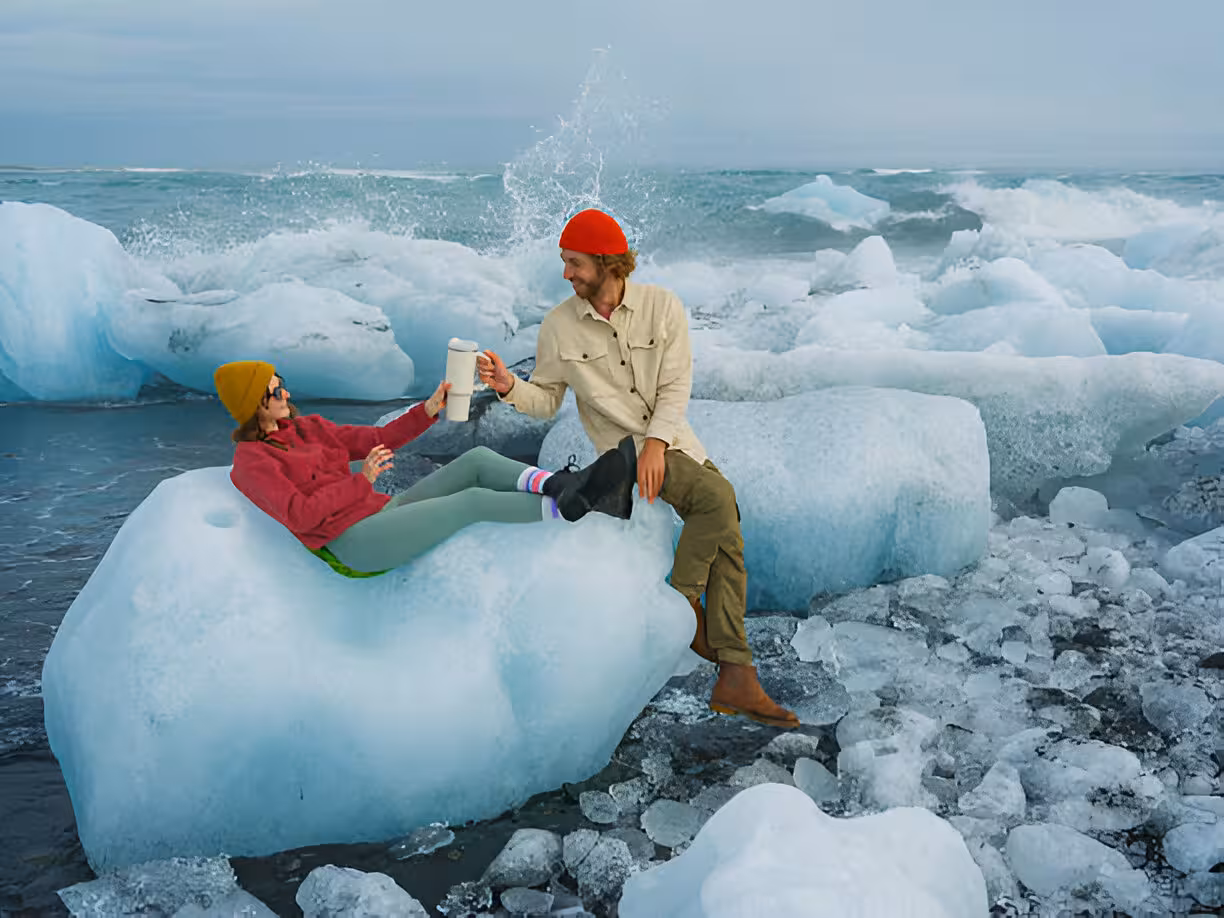 Couple sitting on ice blocks at Diamond Beach Iceland with coffee cups and ocean waves
