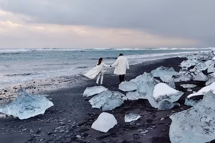 Couple in white coats holding hands on Diamond Beach black sand shore with glacier ice chunks