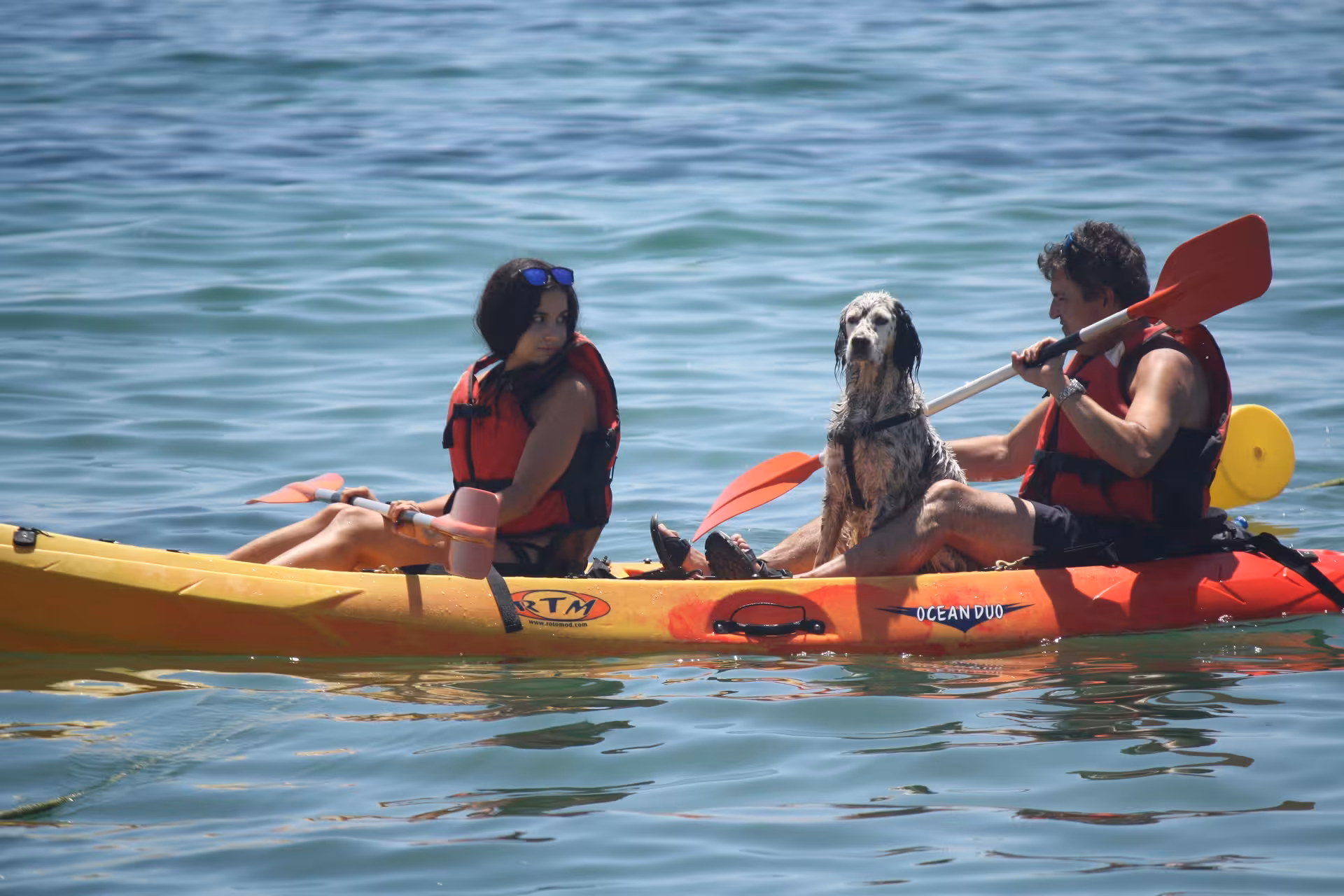 Couple with their dog enjoying a tandem kayak tour on calm blue sea wearing red life jackets