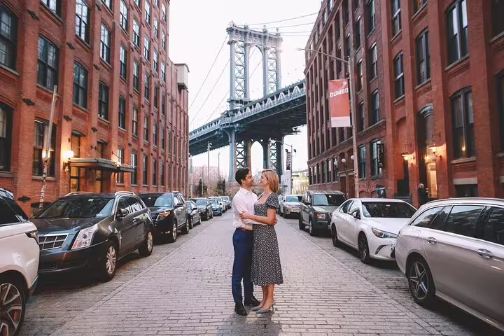Couple embraces on cobblestone street in DUMBO, Brooklyn, with the iconic Manhattan Bridge towering above them.