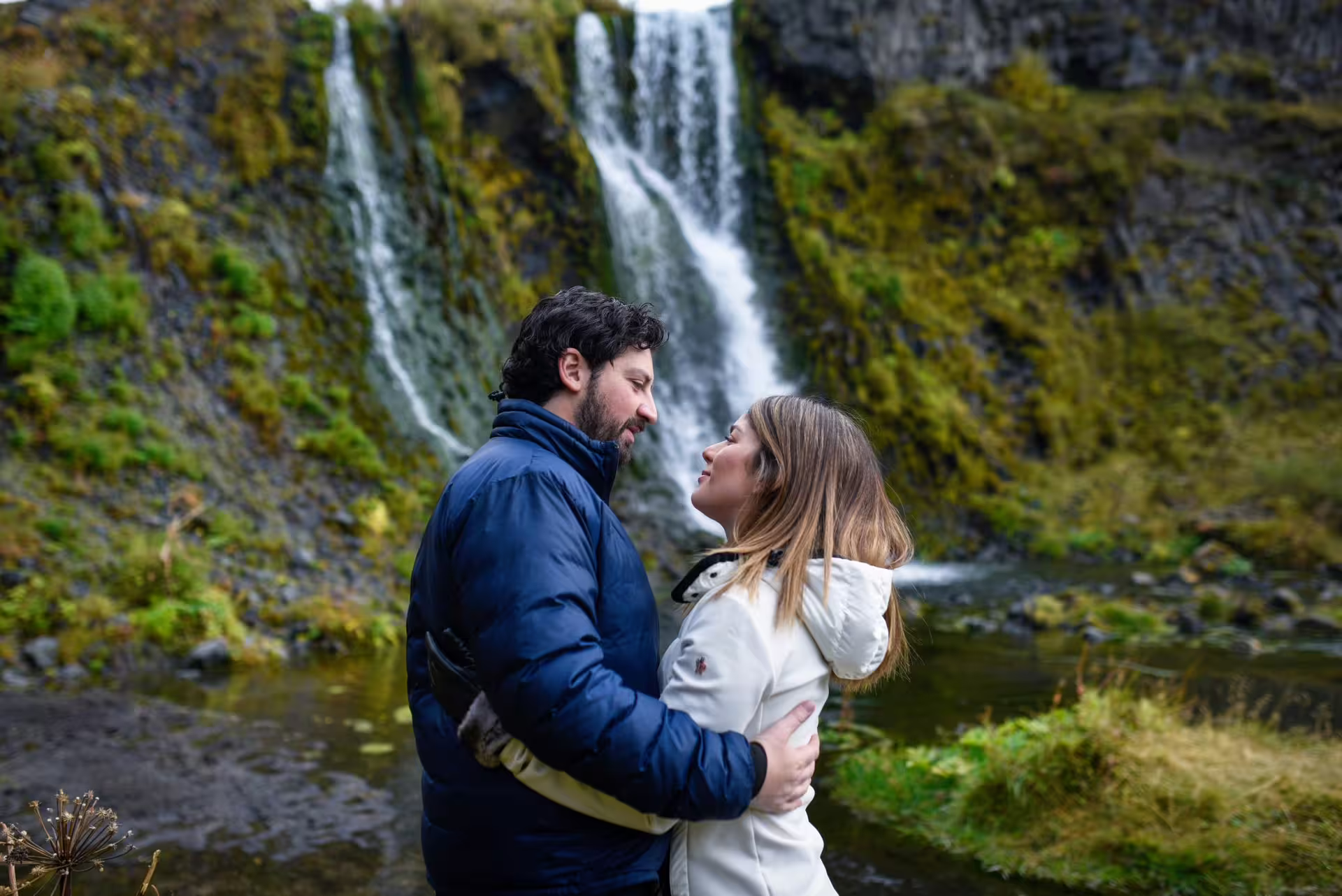 A romantic couple embraces in front of a cascading Iceland waterfall during an unforgettable 12-day grand tour.