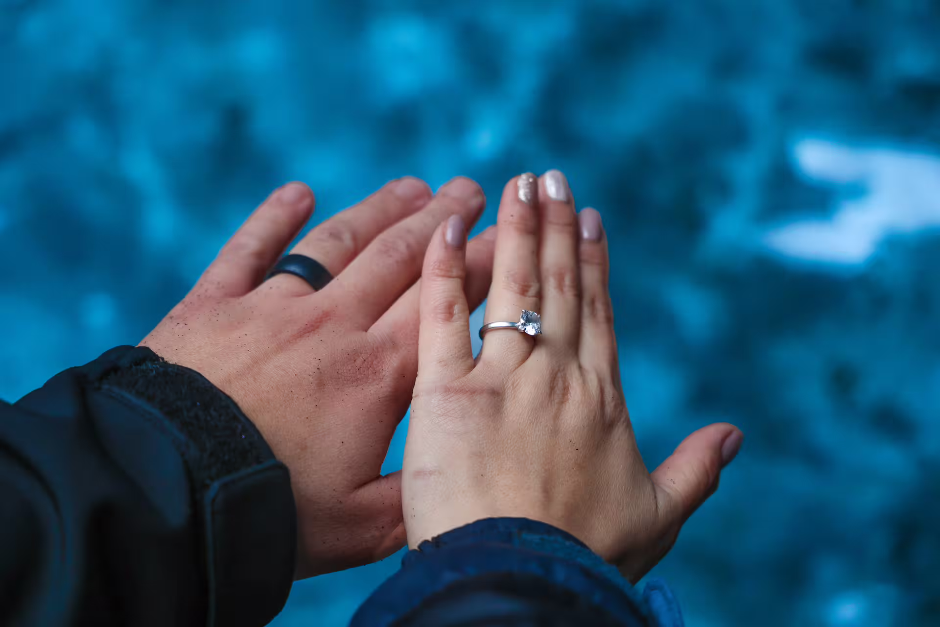 Close-up of couple's hands showing engagement ring against glowing blue glacier backdrop after proposal